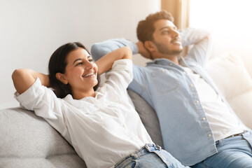 Indian couple leisurely reclining on sofa, holding hands behind heads