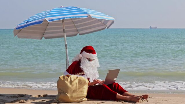 Santa Claus sits under a beach umbrella against the backdrop of the sea on a hot summer day and types something on a laptop, looking into a bag of gifts. Santa's summer vacation.