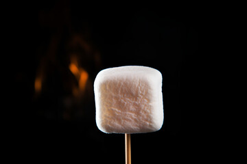 Single marshmallow on a wooden stick over dark background with a hint of fire, copy space. Grilled marshmallow skewer. Cooking skewers with grilled marshmallows. Close up of grilling marshmallows
