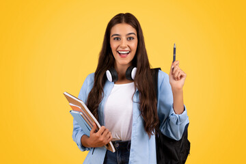 Cheerful european woman student, with books and headphones, rise pen up, got idea sign