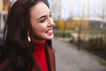 Profile of a young woman in a warm autumn coat, smiling sincerely while walking down the street.