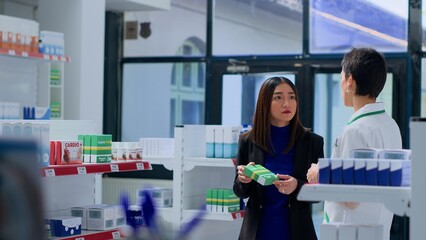 Obraz premium Asian woman in dispensary needing prescription drug to fight off virus, asking druggist to help her pick best medicament. Pharmacy technician assisting customer with product sugestion