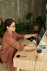 Side view of young female photographer adjusting computer monitor while sitting by workplace in studio and making review of new shots
