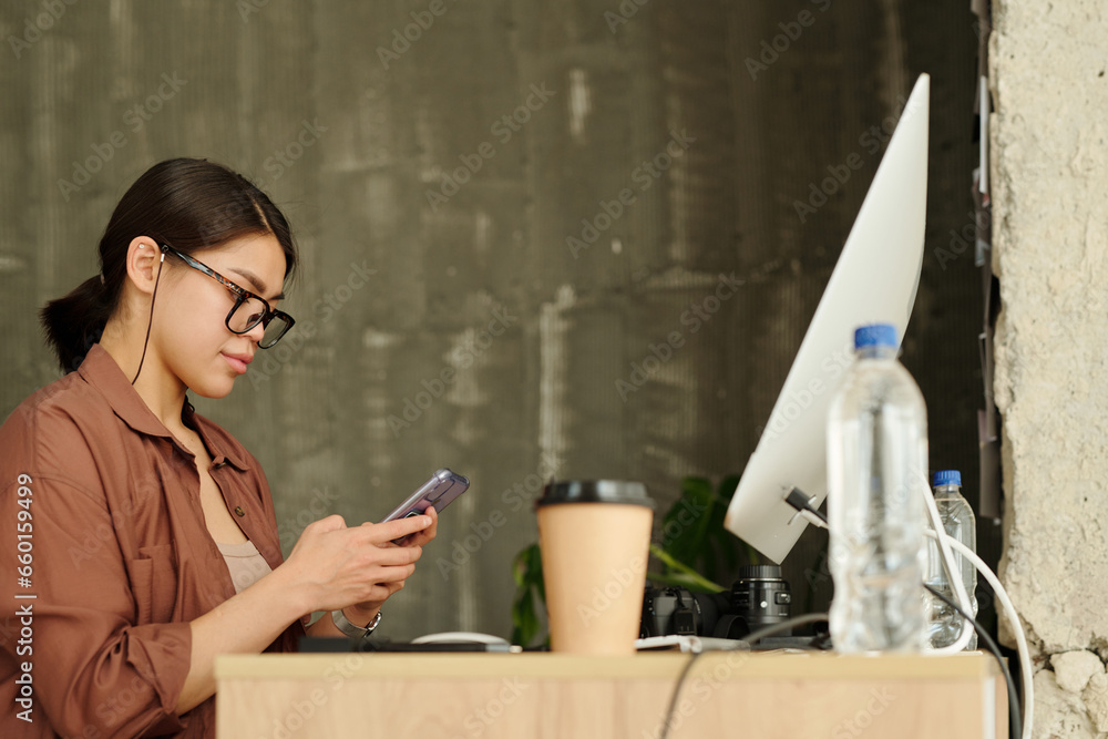 Aufkleber young modern female designer or photographer texting in smartphone while sitting by workplace in fro