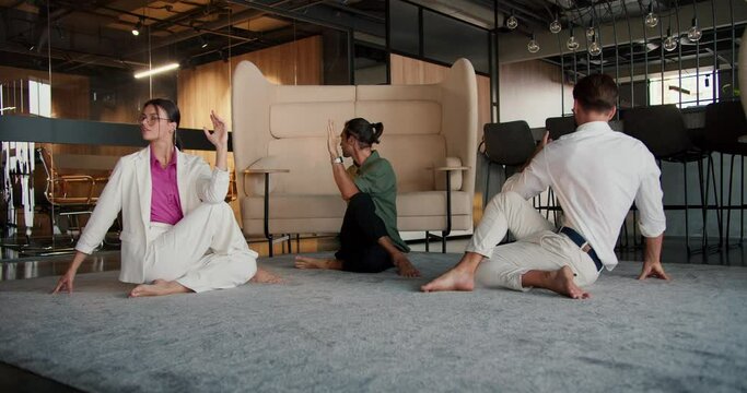 A girl and two guys in white and green business suits are sitting on a rug in the office and doing a Spine Twisting Pose. Yoga classes during a break from office work