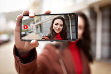 A young woman on the street is video blogging on her phone, her image visible on the phone's screen. video blogging concept