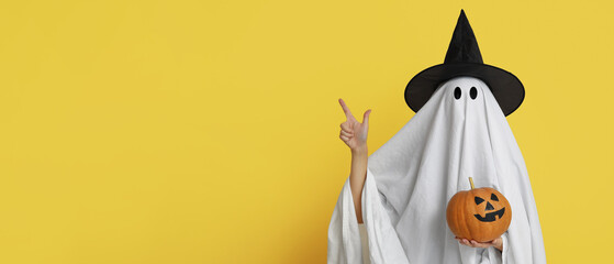 Person in costume of ghost holding Halloween pumpkin and pointing at something on yellow background...