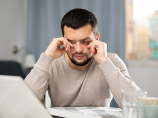 Man rubs tired eyes with his hands after a long work at a laptop at home