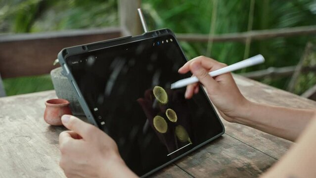 Female artist hands drawing with stylus on tablet at the cafe on tropical island, palm trees. Young designer works remotely open air at exotic location. Pretty woman editing illustration. Close-up.