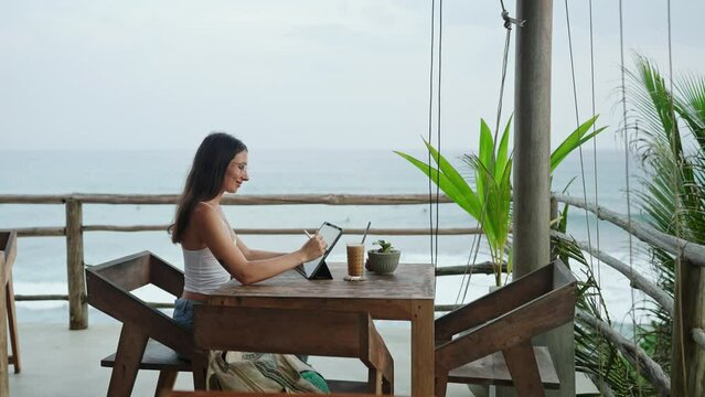 Young illustrator drawing on tablet at cafe with ocean view, palm trees. Pretty focused woman works remotely at authentic bar near tropical coast. Freelancer editing illustration at the seaside.