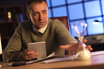 Shocked mature man with tablet computer and hourglass in office at night