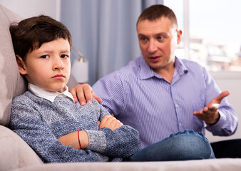 Young father consoling preteen son sitting at sofa after quarrel at home