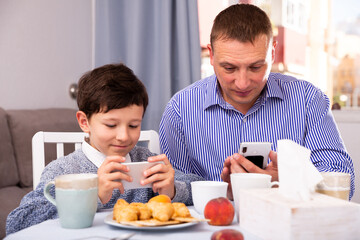 Happy tween boy spending time with his father at home, carried away with mobile gadgets during breakfast