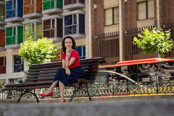 Pretty woman sitting on a bench wearing a red sweater black skirt looking at the camera in the city of Bilbao.
