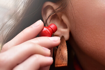 A woman holds a red wireless earphone in her hand, bringing it closer to her ear to listen to music.