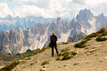 Rear view of male tourist with knapsack on shoulders and trekking pole in hands standing on mountain slope, enjoying scenic view of rugged rocky peaks of Dolomites under cloudy sky