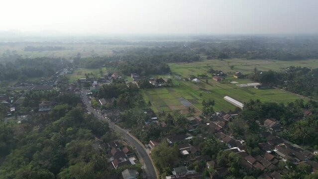 Aerial view of human settlement buildings expansion near paddy rice fields in the morning sunlight. Kulon Progo rural village landscape. Population density is displacing agricultural land and forests.