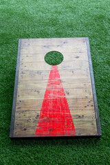 A worn wooden square corn hole board game with a red arrow leading to a small open hole for...