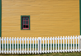 Obraz premium The exterior yellow wooden wall of a vintage building with a small glass window. The window has red and green trim around its six panes. There's a white wooden picket fence in the foreground property.