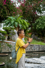 An elderly woman in a yellow T-shirt with a backpack behind her takes a photo with her phone while standing on the rocks next to the pond.