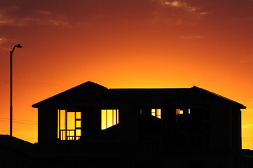 Framed two-story house under construction silhouetted at sunset, showing exposed studs and trusses in residential development phase with street light and power lines