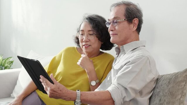 Happiness senior couple sitting on sofa using tablet while video call online with family in living room at home, happy elderly man and woman conversation distant with family with enjoy and relaxation.
