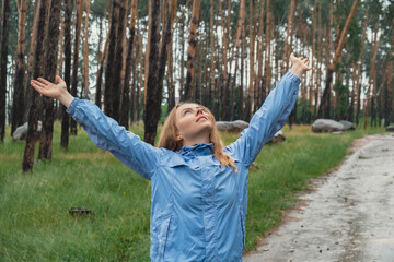 Overjoyed blonde woman in blue raincoat enjoying silence natural green environment woods in the forest. Happy emotion open arms outdoors in rainy weather. Unity with nature physical mental wellbeing 