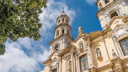  An old Catholic cathedral in the historic quarter of Buenos Aires. There are columns, carved ornaments, sculptures on the facade. A tall tower with a bell and a cross,  blue sky and clouds. Argentina. © Вера 