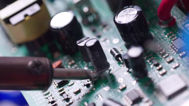 Close-up of a technician soldering a capacitor to a circuit board with a wisp of smoky fumes coming out of it.