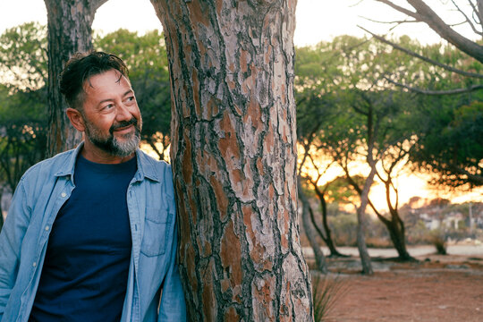 One man  standing against a tree in the park with serene and smile expression looking on the side. People enjoy weekend activity and outdoor leisure moment. Casual denim clothes. Modern senior male