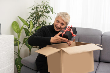 Positive middle aged older woman sitting on couch with carton box on laps, feeling curious about ordered wished item from website, smiling female client satisfied with fast courier delivery service.