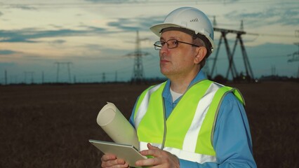 Engineer with blueprint works on tablet and smiles at power substation