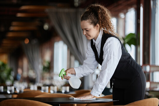 Side view portrait of smiling young woman as server wearing classic uniform and cleaning dining tables in restaurant, copy space