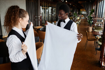 Portrait of two smiling elegant servers folding white tablecloth while preparing luxury restaurant...