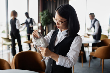 Waist up portrait of Asian young woman as classic server polishing glasses in luxury restaurant, copy space