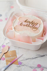 Small birthday cake in a white gift box with a spoon and candle. Trendy Korean style bento cake with Happy birthday inscription and pink pastel cream smears on top.