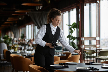 Portrait of young woman as female server setting tables in luxury restaurant preparing for opening,...