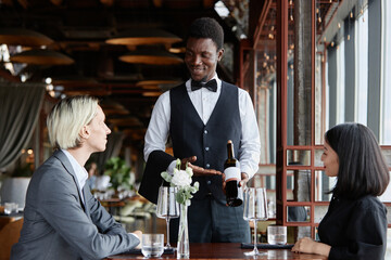 Portrait of Black young man as server taking orders from couple in luxury restaurant and presenting bottle of red wine, copy space