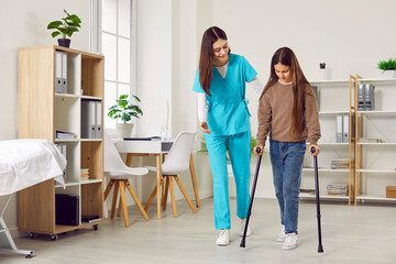 Woman doctor or nurse in a medical scrubs uniform helps a little kid child patient girl walk with...