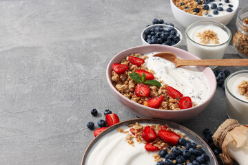 Bowls with yogurt and berries, wooden spoon and milk bottle on gray background, space for text