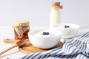 Bowls with yogurt, kitchen board, wooden spoon and bottles on white background