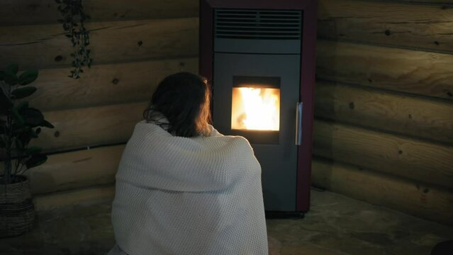 A young Caucasian woman is sitting by an electric pellet fireplace wrapped in a blanket.