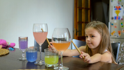 Little girl, toddler plays music with drumsticks in glasses of water in the kitchen