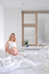 A young blonde woman waiting for a baby is sitting on a bed in a white Scandinavian interior. Pregnancy.