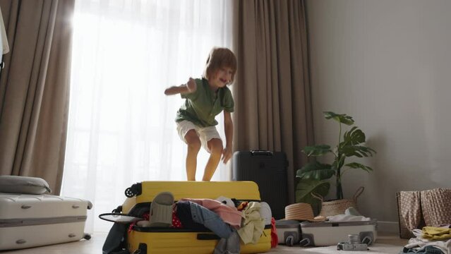 Child jumps on packed suitcase, rejoices, smiles. Little boy, mischievous troublemaker, made mess in packed suitcase and tossed all clothes. Boy jumps on top of suitcase and dances contentedly happily