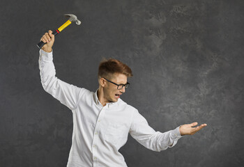 Crazy angry young man holding hammer over something imaginary holding in his hand. Caucasian emotional man who is stressed or has problems. Concept of anger, frustration and burnout. Gray background.