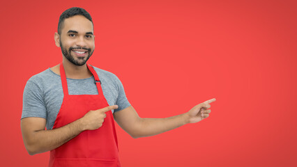 Laughing hispanic waiter with apron pointing sideways on red background