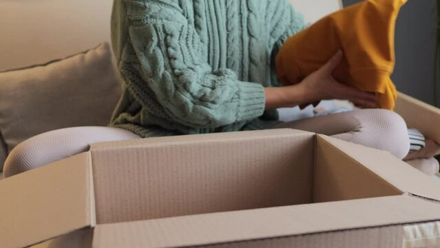 Unrecognizable woman in knitted shirt sitting on couch in home interior packing her attires to carton parcel preparing for sending to poor people donation volunteering.