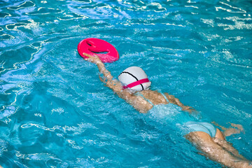 Kid girl swimmer in swimming white cap training with float in swim pool, learning. Child five year...