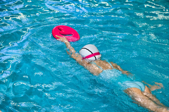 Kid girl swimmer in swimming white cap training with float in swim pool, learning. Child five year old in swimsuit and swimming goggles exercising in pool. Workout learn concept. Copy ad text space